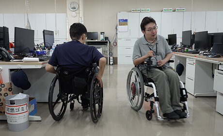 デスクが並ぶオフィスの広い通路で、車いすに乗った社員2人がすれ違っている様子