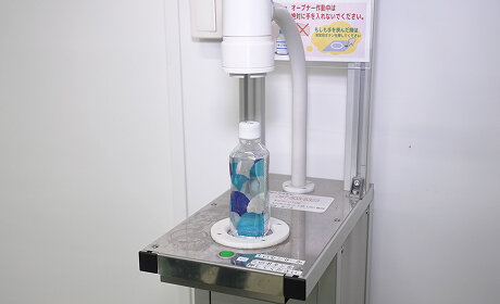 台座にペットボトルを置くと、上部の装置がキャップを自動で開ける自動キャップオープナー。「オープナー作動中は絶対に手を入れないでください。」の注意書きが貼られている