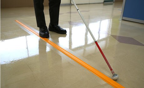 通路の床に貼られた黄色のココテープに沿って、白杖を持った人が歩いている足元の写真