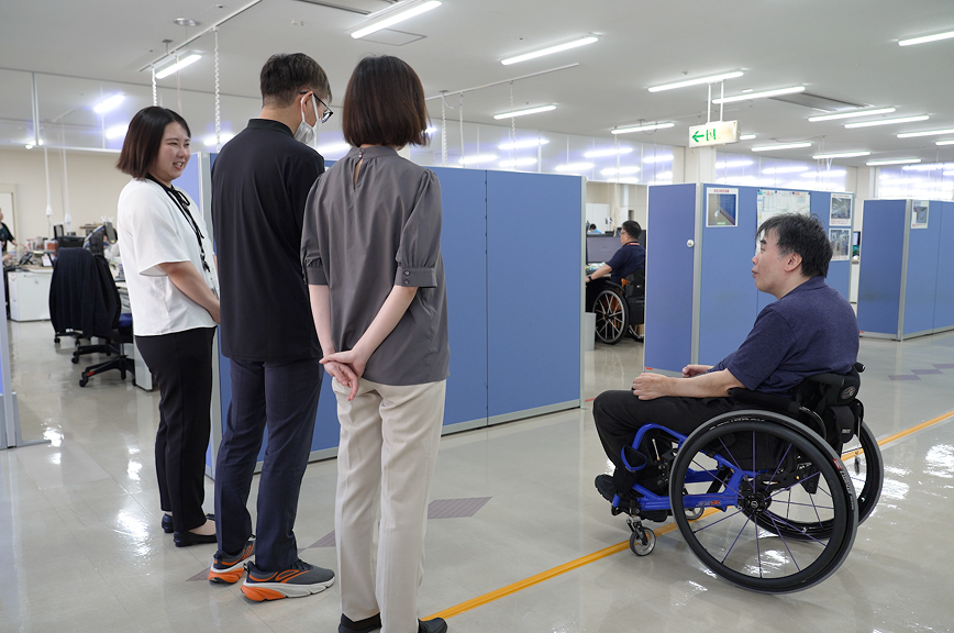 青いパーテーションが並ぶオフィスの通路で、車いすに乗った社員が、立っている3人の見学者に説明している様子