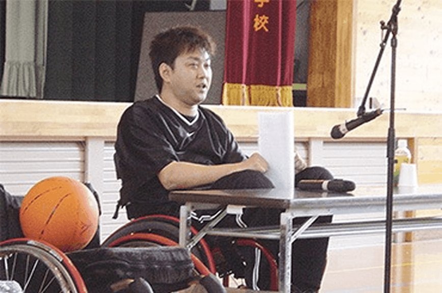 学校の体育館で、車いすに座った社員がマイクの前で講演している様子